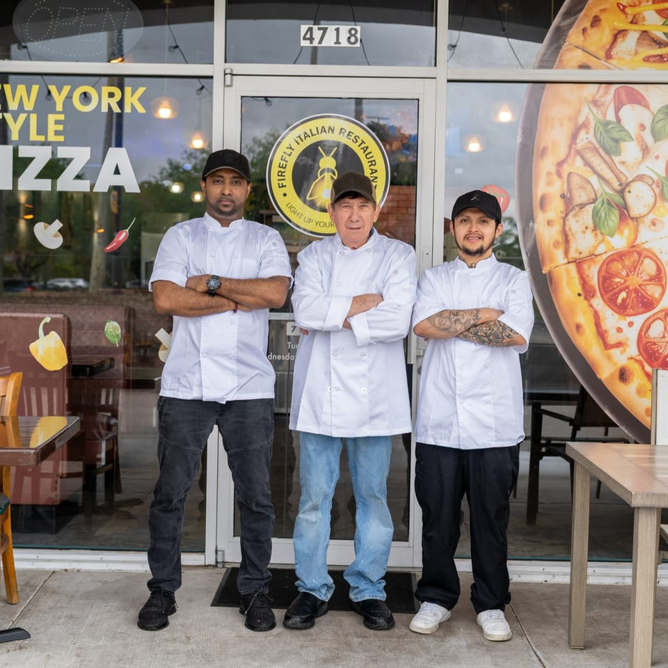 Three chefs in white coats and hats stand with crossed arms in front of Firefly Italian Restaurant.
