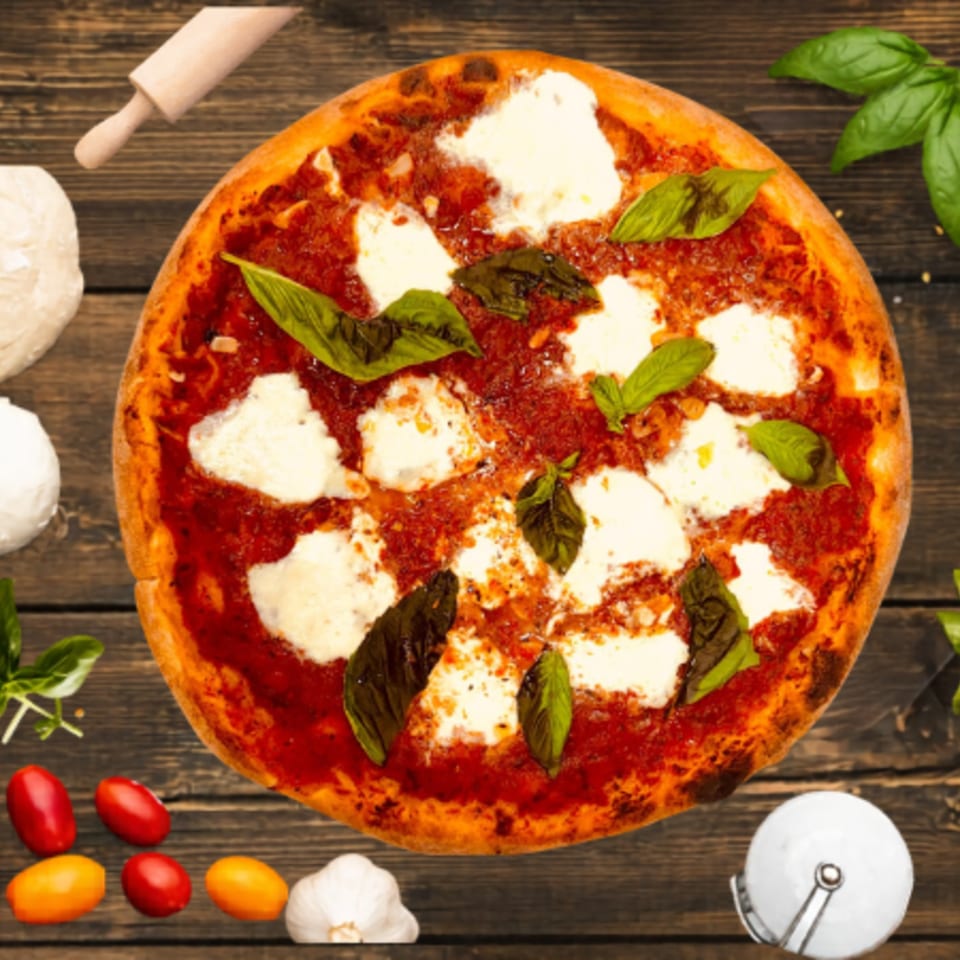 A freshly baked Margherita pizza with basil leaves, tomatoes, garlic, and a rolling pin on a wooden table.