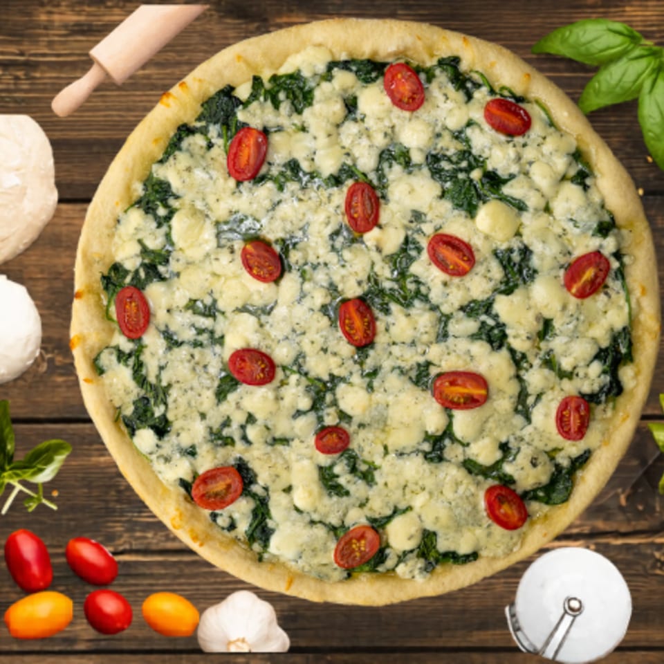A white pizza with spinach, cheese, and cherry tomatoes on a wooden table with ingredients.