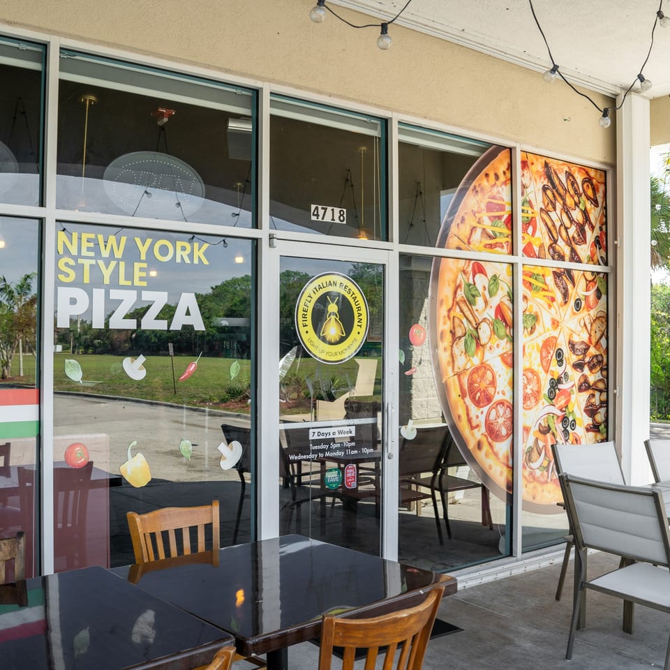 Exterior of Firefly Italian Restaurant with "New York Style Pizza" on windows and outdoor seating.
