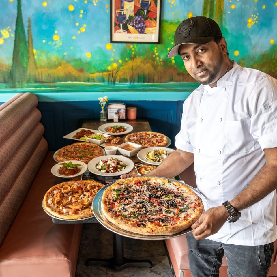 Chef in a white coat and black cap holding a pizza, with a table full of Italian dishes.