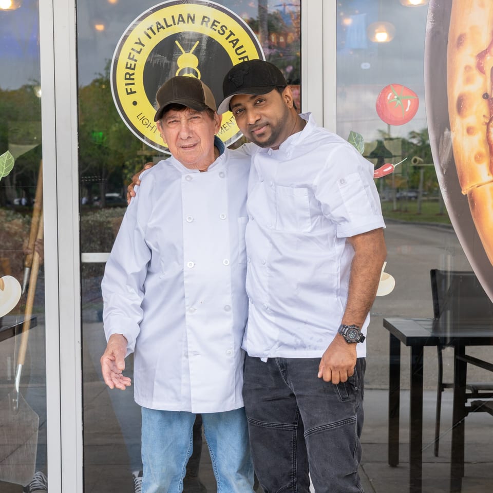 Two chefs, one older and one younger, stand smiling in front of the Firefly Italian Restaurant entrance.