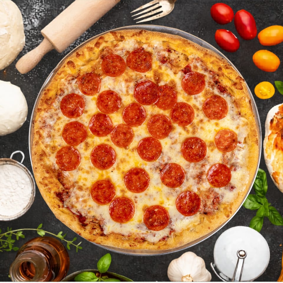 A pepperoni pizza with melted cheese, surrounded by fresh ingredients like tomatoes, garlic, and basil.